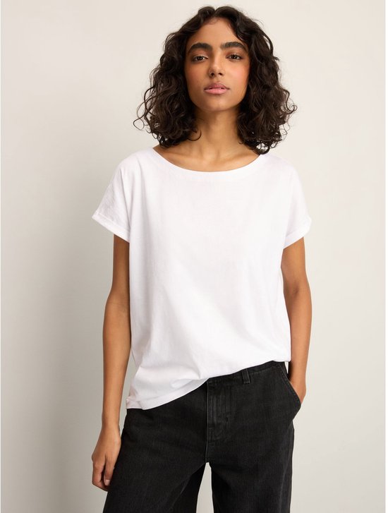 Een vrouw met krullend donker haar draagt een wit T-shirt met opgerolde korte mouwen en een ronde hals, met haar handen in de zakken.