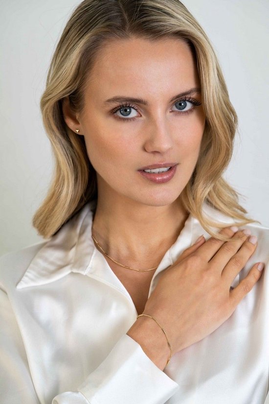 Model met blonde haren draagt een gouden ketting en armband, witte blouse.