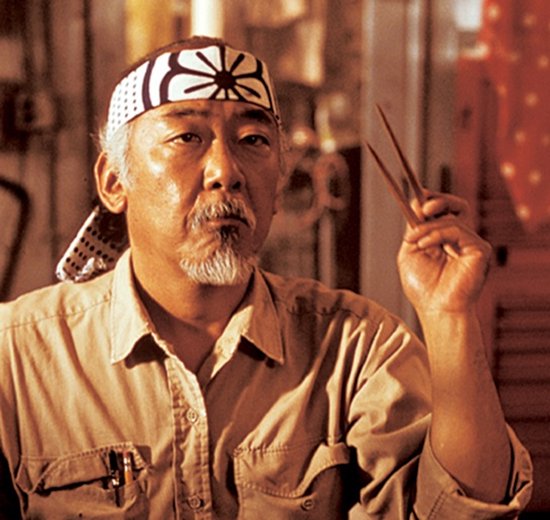 Een close-up van de oude man Mr. Miyagi (Pat Morita) uit Karate Kid met een witte band om zijn hoofd en een pincet in zijn hand.