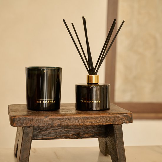 Zwarte glazen diffuser en zwarte kaars van Ted Sparks met gouden details, op een houten krukje.