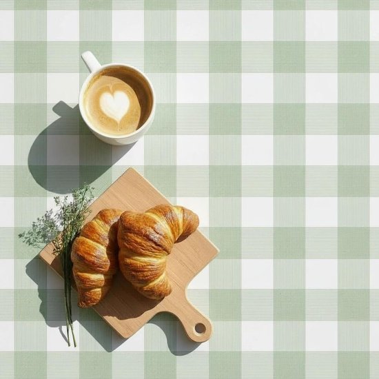 Ontbijttafel gedekt met groen en wit geruit tafelzeil, met koffie en croissants.