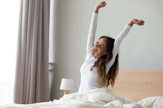 Een glimlachende vrouw met lang haar, in een wit shirt, die zich uitrekt in bed.