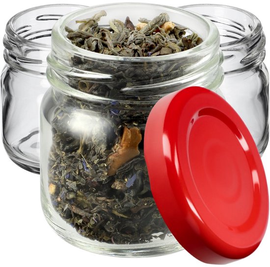 Een glazen potje met groene thee, een rood deksel ernaast en twee lege glazen potjes erachter, op een witte ondergrond.