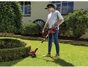 Vrouw in grijze top en spijkerbroek gebruikt rode grastrimmer in een tuin met gesnoeide struiken.