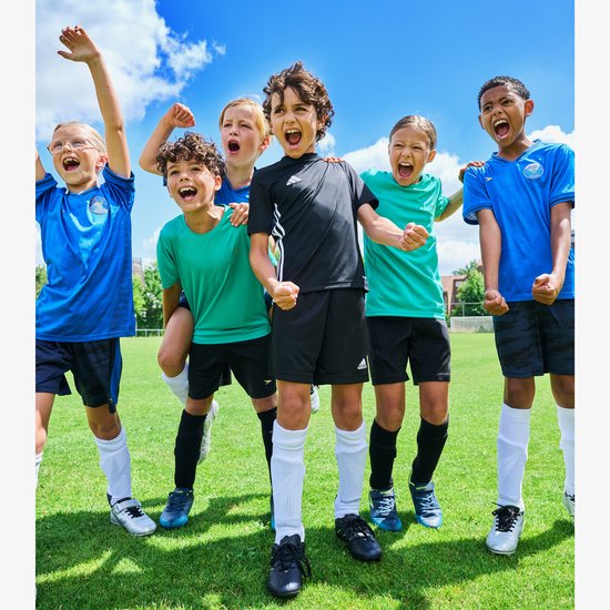 Een groep lachende kinderen in zwarte, groene en blauwe voetbaltenues op een grasveld, sommigen juichen.