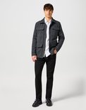 Man met grijze jas, wit overhemd en zwarte jeans, staand.