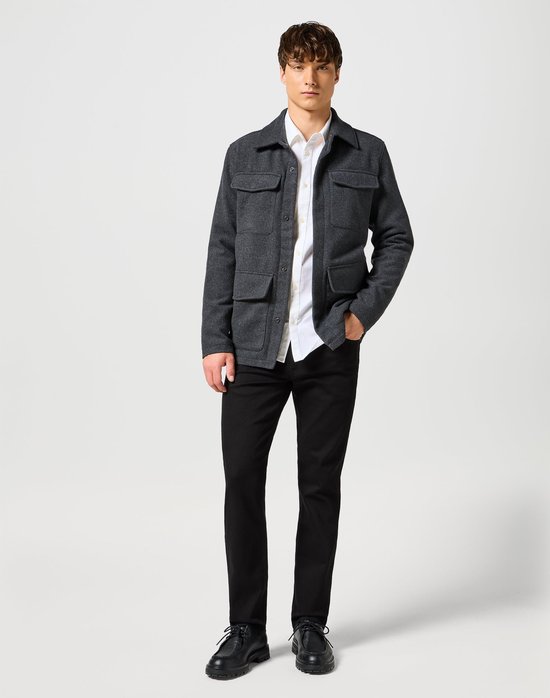 Man met grijze jas, wit overhemd en zwarte jeans, staand.