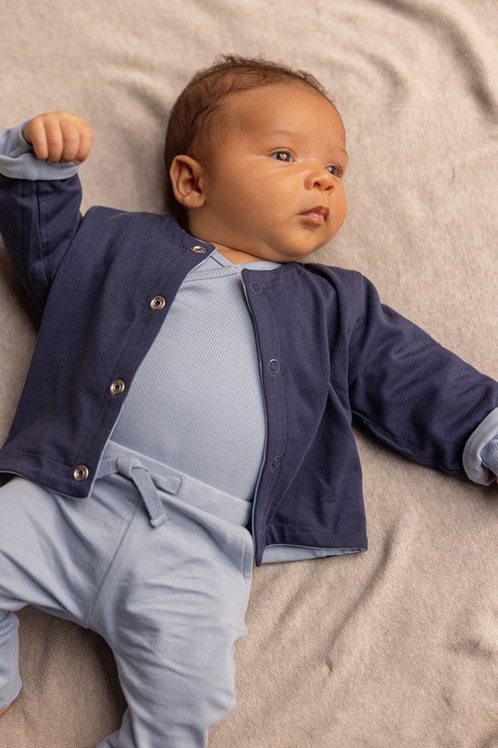 Baby in een lichtblauw tenue, bestaande uit een body en een joggingbroek, met een donkerblauw vestje.