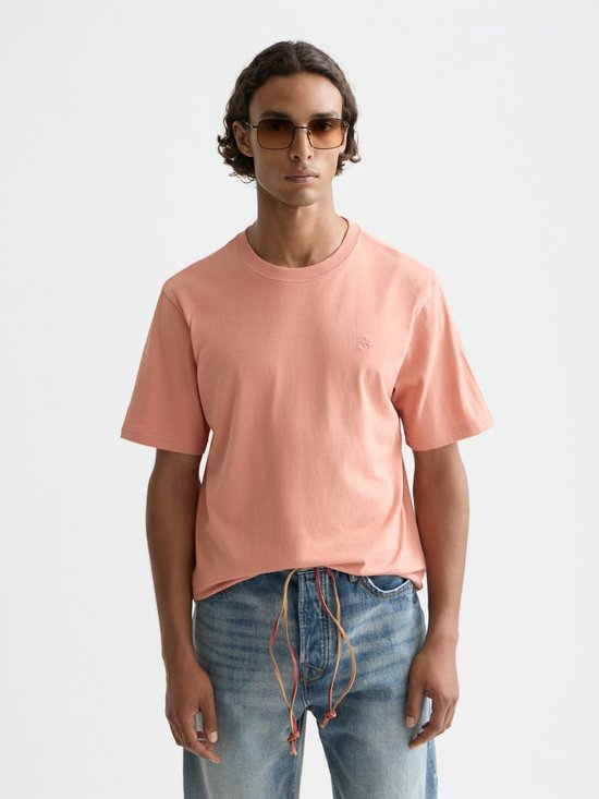 Model draagt een perzikkleurig T-shirt met korte mouwen en jeans, zonnebril op.