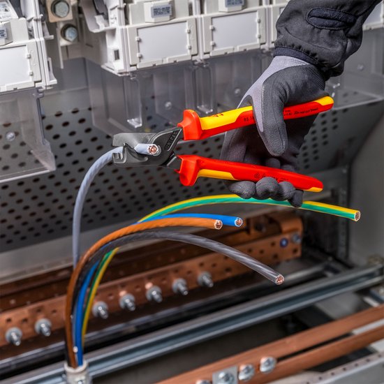 Knipex VDE stap-snij XL kabelschaar in gebruik bij het knippen van een grijze kabel in een schakelkast, met gekleurde kabels eronder.