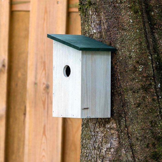 Grijs houten vogelhuisje met schuin groen dak, gemonteerd op een boomstam voor een houten schutting.