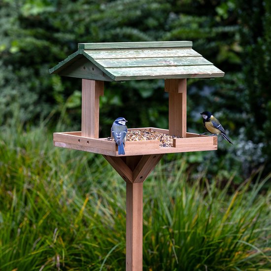 Houten vogelhuisje op paal met een groen dak, bezocht door twee kleine vogels op een plateau met vogelzaad in een tuin.