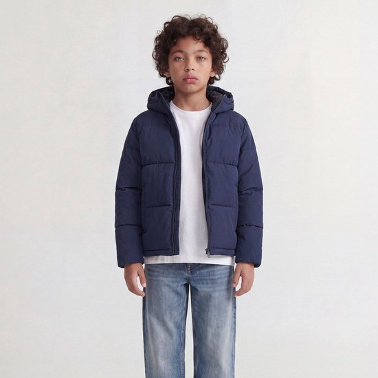 Jongen in een donkerblauwe, gewatteerde jas met capuchon, witte T-shirt en jeans.