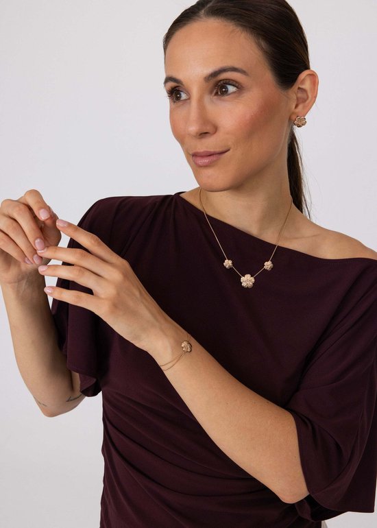 Model draagt gouden Purelei ketting, oorbellen en armband met bloemmotief. De sieraden passen bij haar elegante outfit.