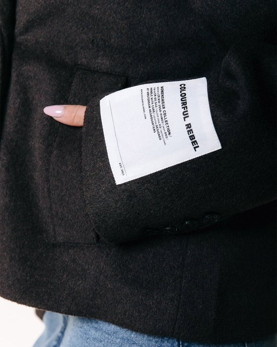 Close-up van de mouw van een donkerbruine wollen jas met een label met de tekst 'COLOURFUL REBEL'.