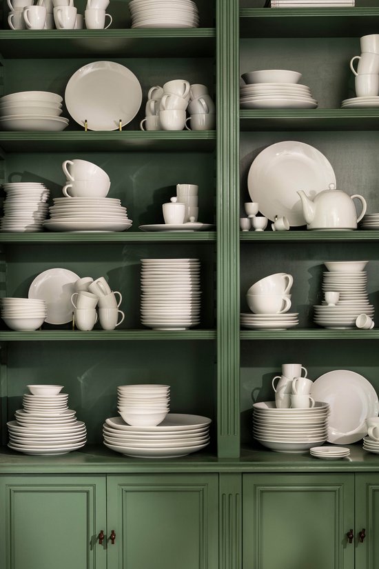 Witte serviezen gestapeld in een groene open kast, met schalen, borden en kopjes.