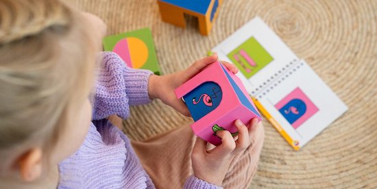 Kind met paarse trui houdt een roze blokje vast met een afbeelding van een flamingo, naast een opdrachtenboekje.