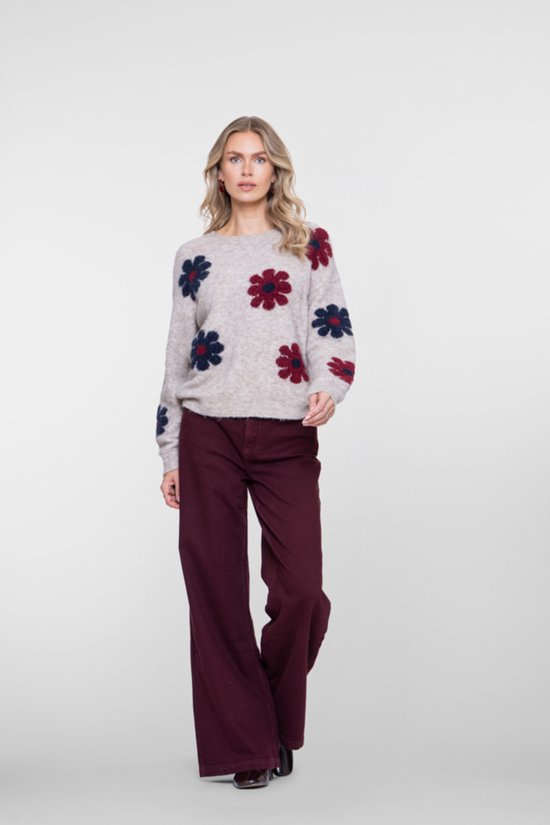Model van voren, draagt een gebreide trui met rode en blauwe bloemen en een donkerrode spijkerbroek met wijde pijpen.