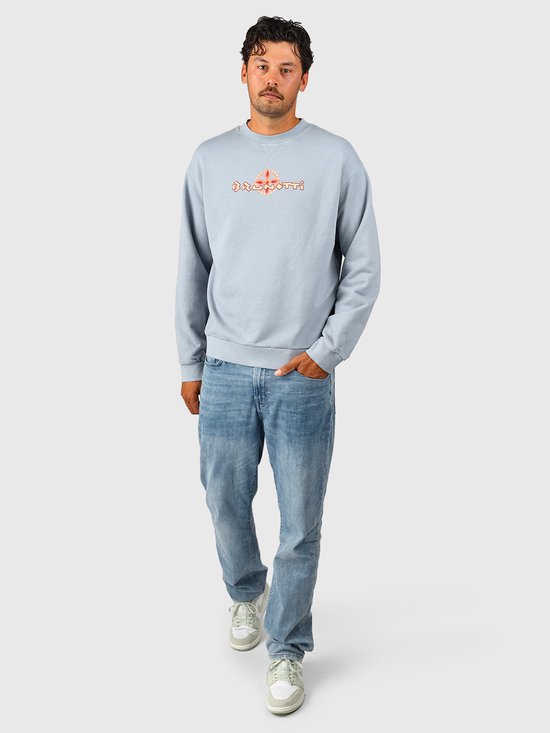 Volledig figuur in lichtblauwe Brunotti sweater en jeans, met oranje-rood logo op de borst.