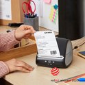 Iemand houdt een geprint Dymo-verzendlabel vast naast een zwarte Dymo LabelWriter 5XL op een houten bureau.