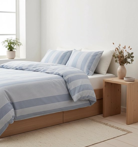Blauw gestreept dekbedovertrek op een bed met houten frame, een plant bij het raam en een vaas met gedroogde bloemen.