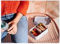 Une femme en jean et chemisier orange tient une bouteille, à côté d'une boîte à lunch grise ouverte avec sandwichs et légumes.