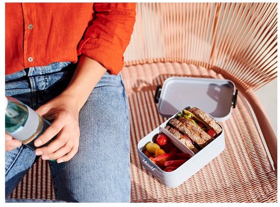 Une femme en jean et chemisier orange tient une bouteille, à côté d'une boîte à lunch grise ouverte avec sandwichs et légumes.
