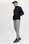 Man in driekwart profiel met zwarte jas, grijze joggingbroek en zwarte sneakers. Witte achtergrond.