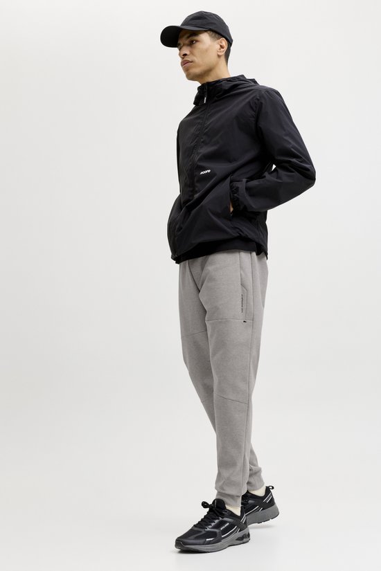 Man in driekwart profiel met zwarte jas, grijze joggingbroek en zwarte sneakers. Witte achtergrond.