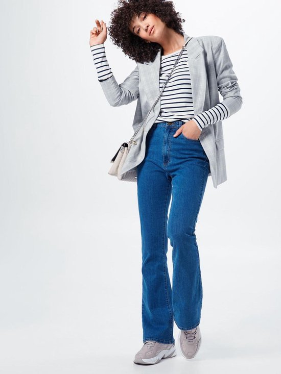 Model met krullend haar draagt een wit gestreept T-shirt met marineblauwe strepen, een grijze geruite blazer, spijkerbroek en een tasje.