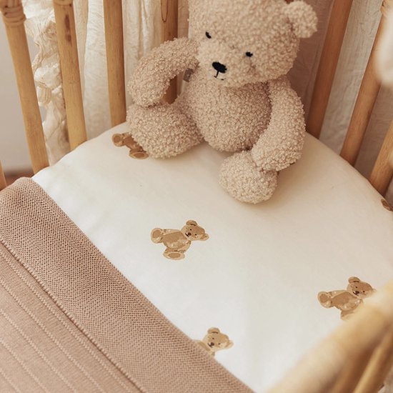 Détail d'un lit de bébé avec un drap de lit crème à imprimé ours en peluche et une couverture tricotée beige.