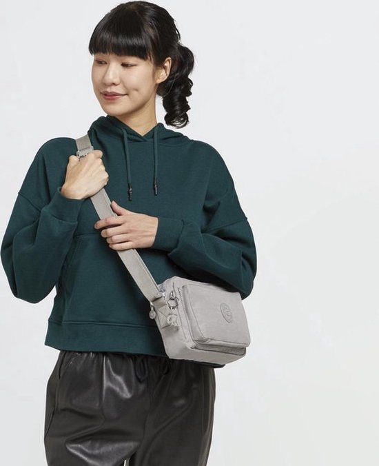 Vrouw met een donkergroene hoodie en zwarte leren broek, draagt een lichtgrijze Kipling schoudertas.
