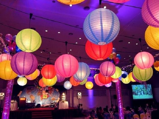 Veelkleurige lampionnen hangen boven een zaal met podium en tafels.
