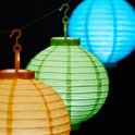 Drie verlichte papieren lampionnen in het oranje, groen en blauw, hangend aan een draad.