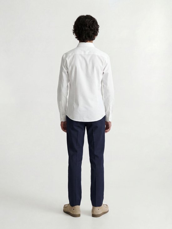 Un garçon porte une chemise blanche à manches longues et un pantalon bleu, vu de dos.