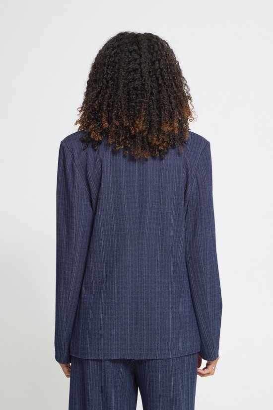 Achterkant van het model met krullend haar, gekleed in een donkerblauwe krijtstreep blazer.