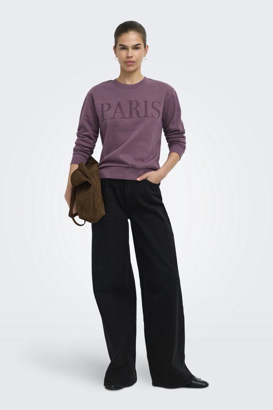Model met paarse sweater met "PARIS" belettering, zwarte wijde jeans en bruine tas.