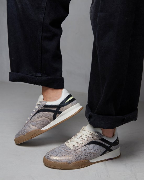 Goudkleurige sneakers met reptielenprint, beige veters en zwarte accenten, gedragen door een persoon in een zwarte broek.