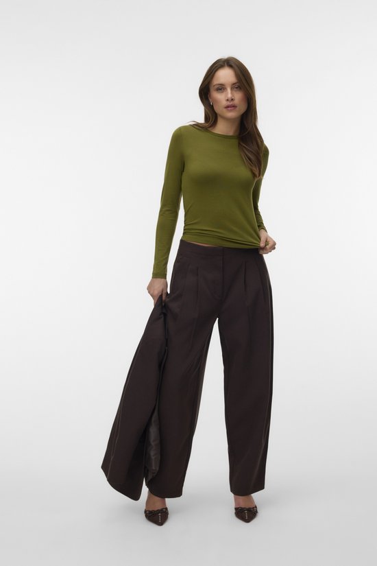 Vrouw staat in een olijfgroen T-shirt met lange mouwen en donkere broek, met jas in haar hand.
