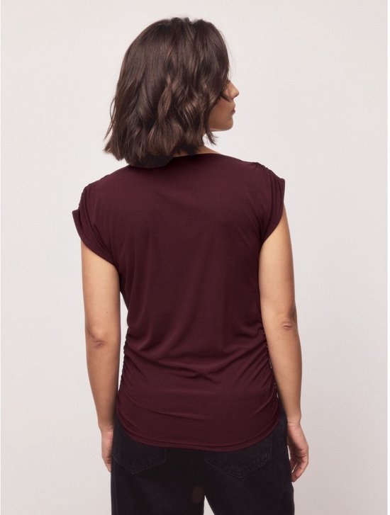 Een vrouw draagt de diepbruine mouwloze top met gedrapeerde hals en zwarte broek, van achteren gezien.