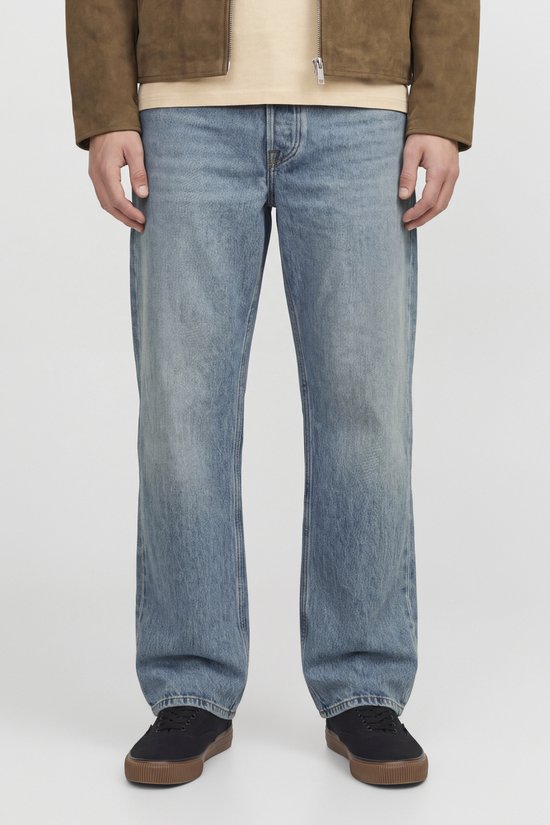 Man in lichtblauwe, wijdvallende jeans, bruine jas, gele trui en zwarte schoenen.