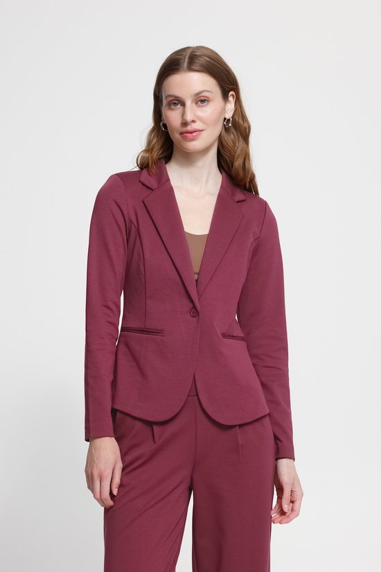Vrouw met lang golvend bruin haar, draagt een bordeauxrode blazer, broek en bruine top. Kijkt naar voren.
