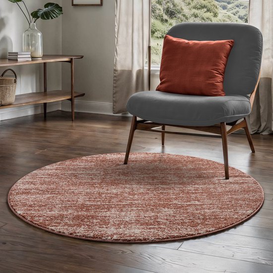 Een terra oranje rond vloerkleed met een subtiel patroon ligt voor een grijze fauteuil met een oranje kussen.