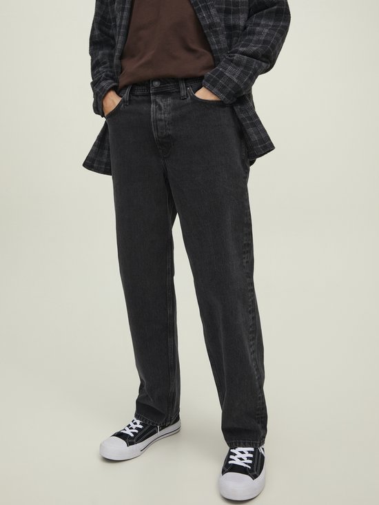 Model in loszittende zwarte jeans en open geruite jas, handen in zakken.