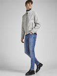 Man kijkt naar links, draagt een grijze jas, zwarte coltrui en blauwe jeans.