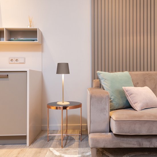 Beige tafellamp met geplooide kap op een koperkleurig tafeltje naast een bank.