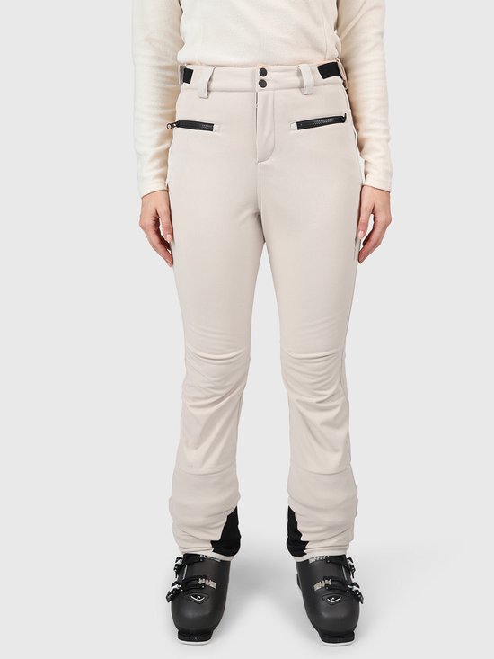 Model in beige skibroek met zwarte ritszakken en ski-uitrusting.