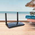 Zwarte TP-Link router met twee antennes, op een houten tafel met boeken en zonnebril bij de zee.