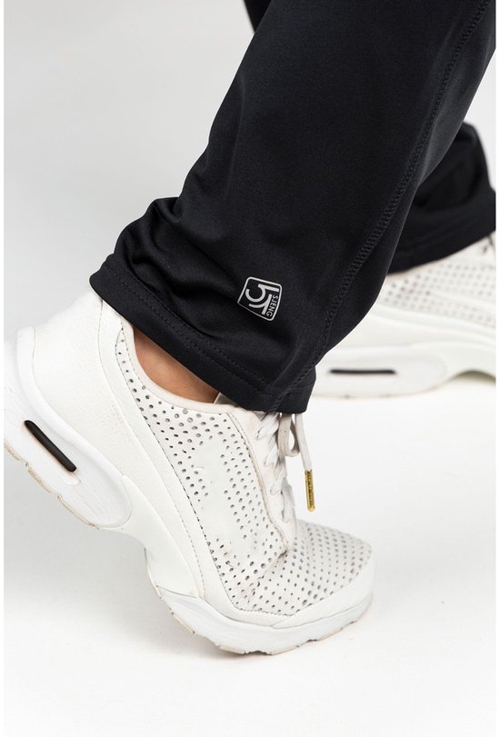 Detail van de onderkant van een zwarte trainingsbroek met het logo en witte sportschoenen.