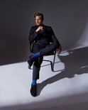Homme en costume bleu foncé et chaussettes bleu vif, assis sur une chaise.